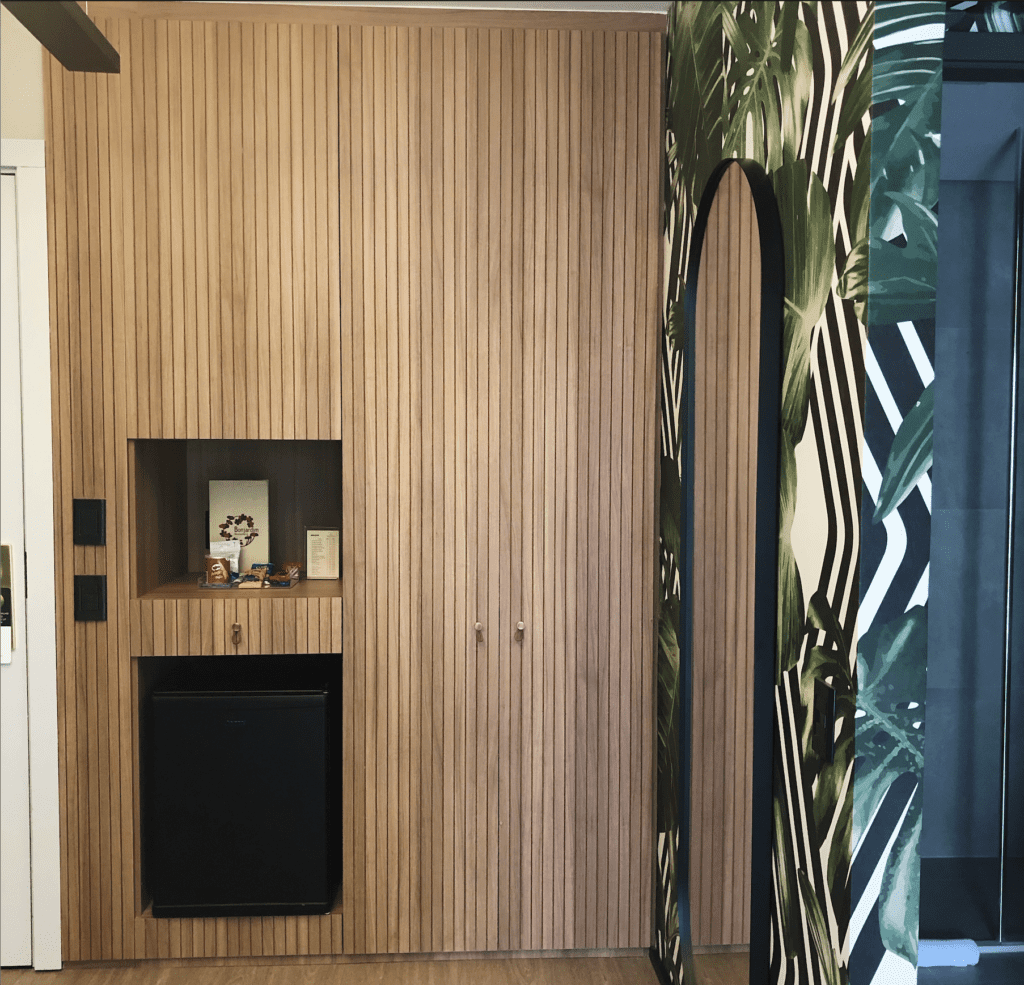 hotel room in Brazil with thin light wood paneling with built in doors and drawers, next to a palm leaf wallpapered wall