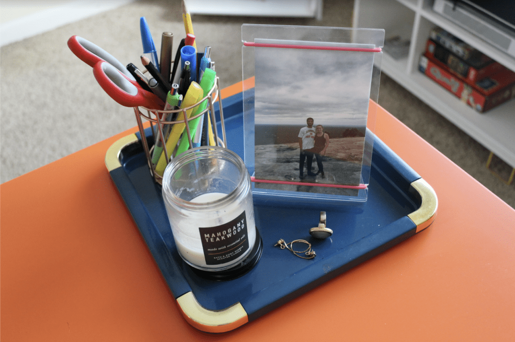 Teal tray on the corner of orange desk with gold pen holder, mahogany teakwood candle, framed photo of girl and man on mountain top, ring, and earrings