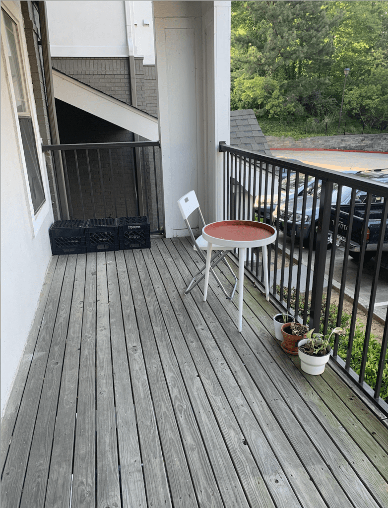 Undecorated balcony with milk crates, folding chair, small orange and white table, and plants on the ground