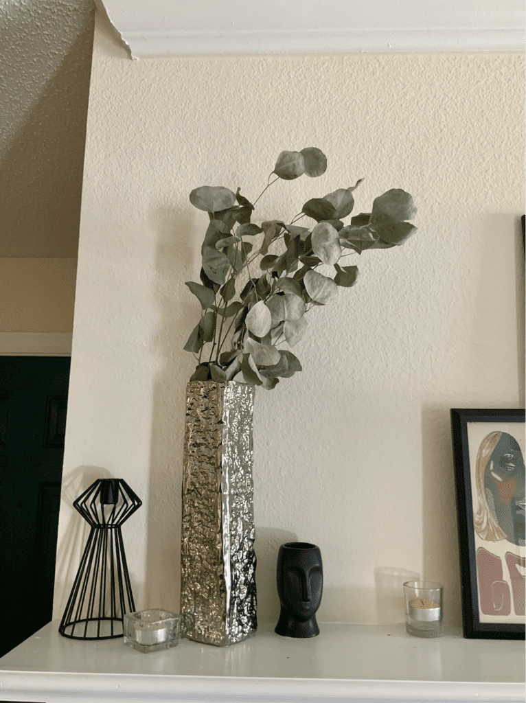 mantel top with thrifted items, including: candle holder, large crushed metal silver vase, and a small empty plant holder in the shape of a face