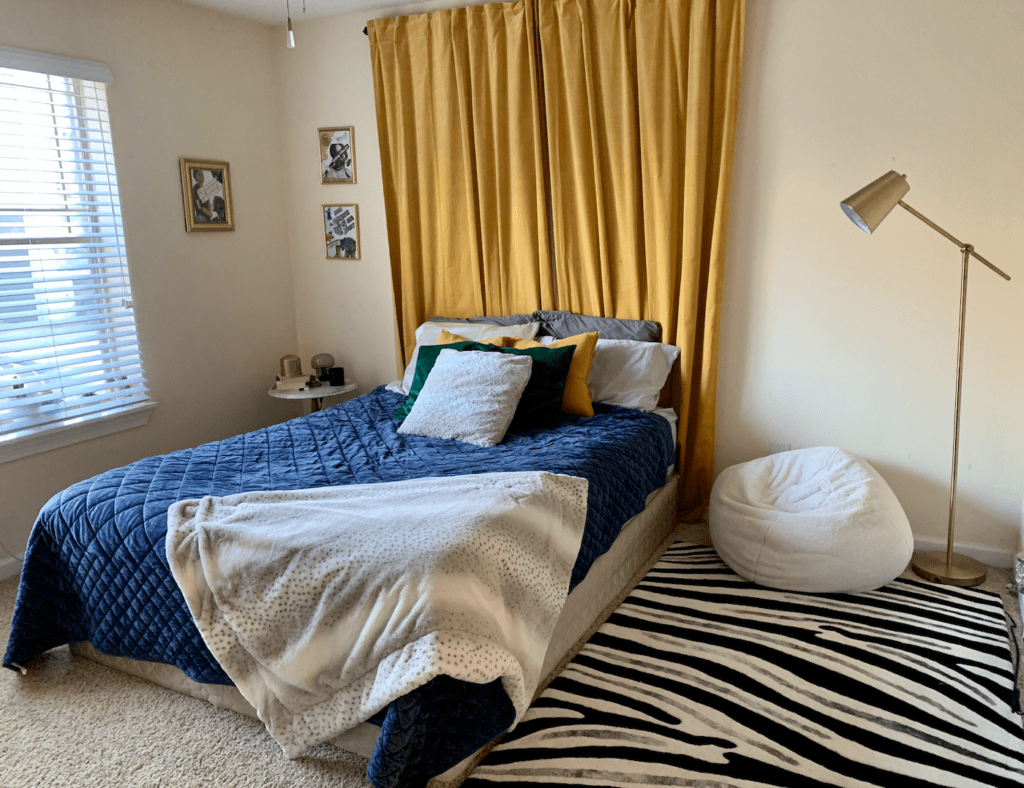 moody jewel tone bedroom with navy blue bedspread, black and white zebra rug, golden velvet curtain hung as a headboard. 
