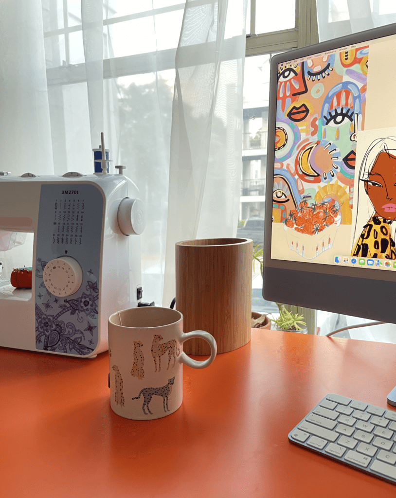 Orange desktop with a blue sewing machine and blue M1 iMac. Small wooden desktop trash can. Coffee cup with colorful cheetahs