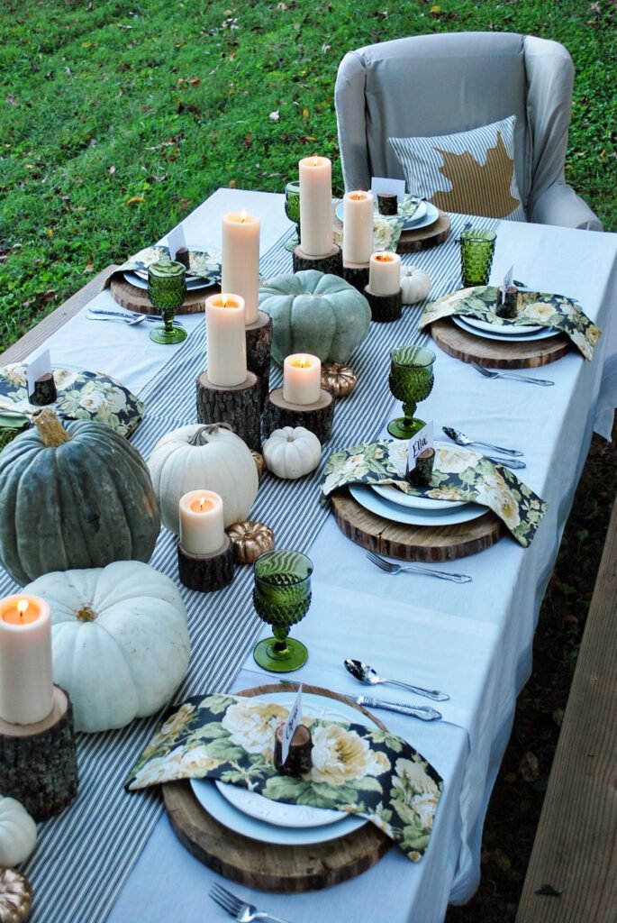 The white buffalo styling co enchanted autumn unique thanksgiving tablescape with wood stump chargers and candle holders, white tablecloth, and white and green pumpkins as a centerpiece
