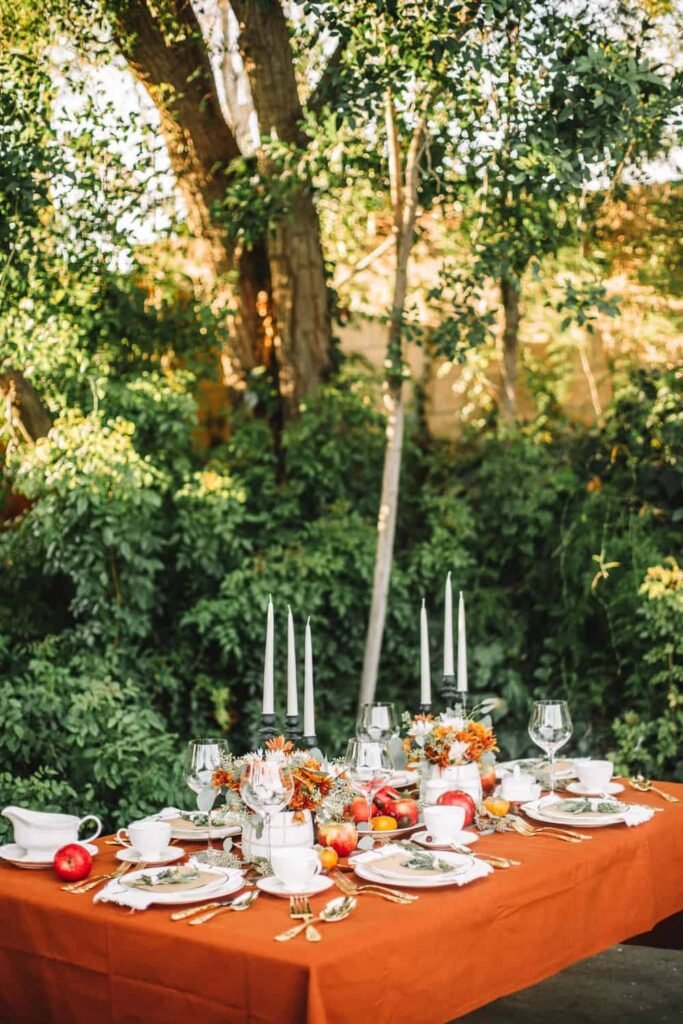 the college housewife modern vintage thanksgiving tablescape with orange tablecloth, orange and red fruit centerpiece, white plates, gold silverware, and black candlesticks