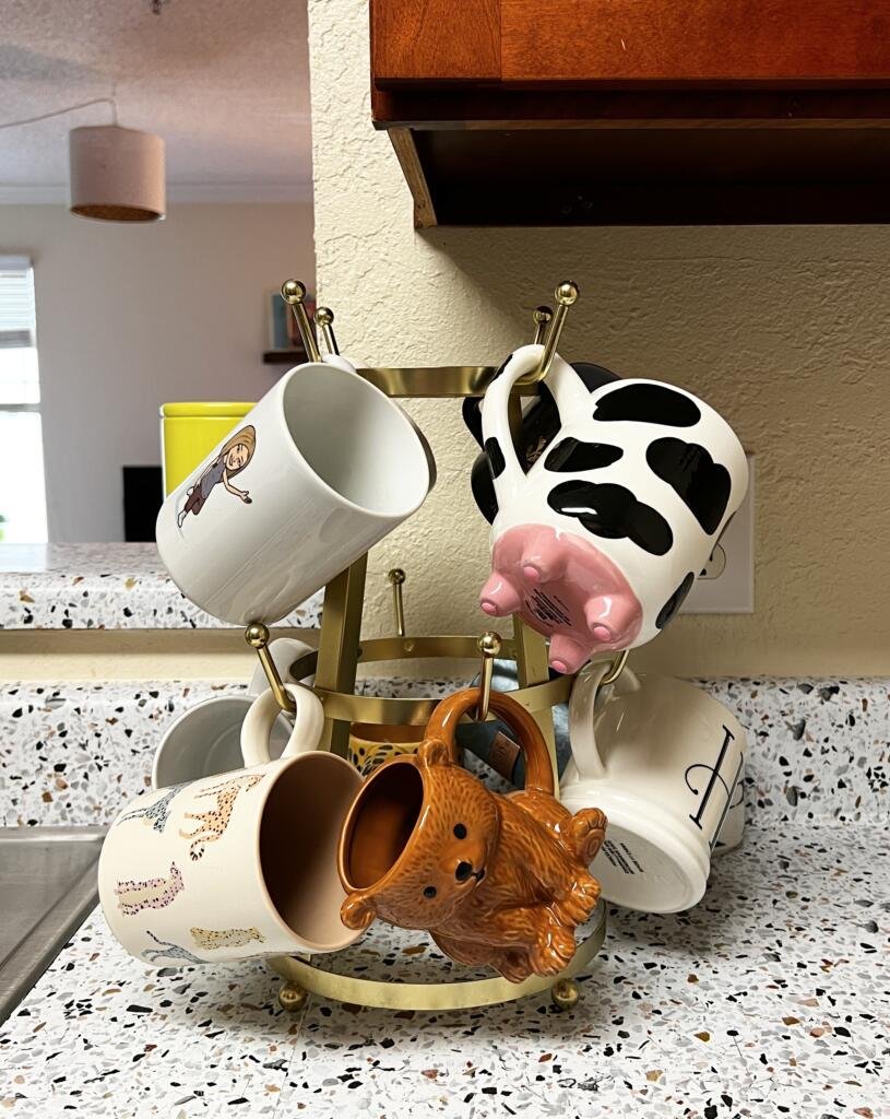 Gold mug rack with a variety of fun mugs full of personality, including a brown bear-shaped mug and a cow mug with pink utters on the bottom