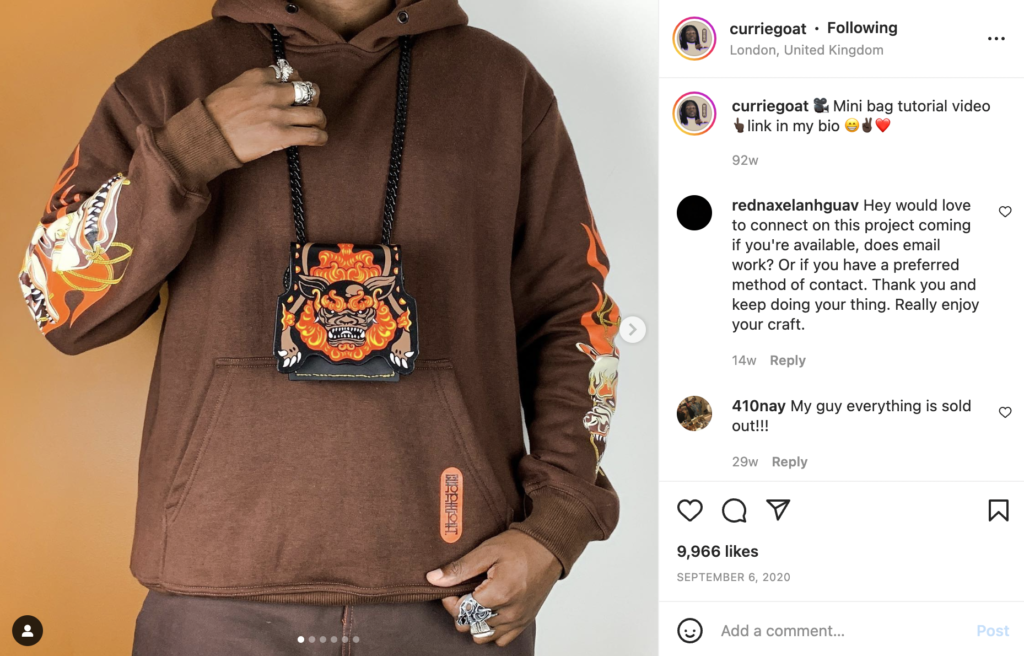 Denzel of curriegoat in a brown hoodie with his design on it, a black, orange, brown and yellow curriegoat bag hanging around his neck. 