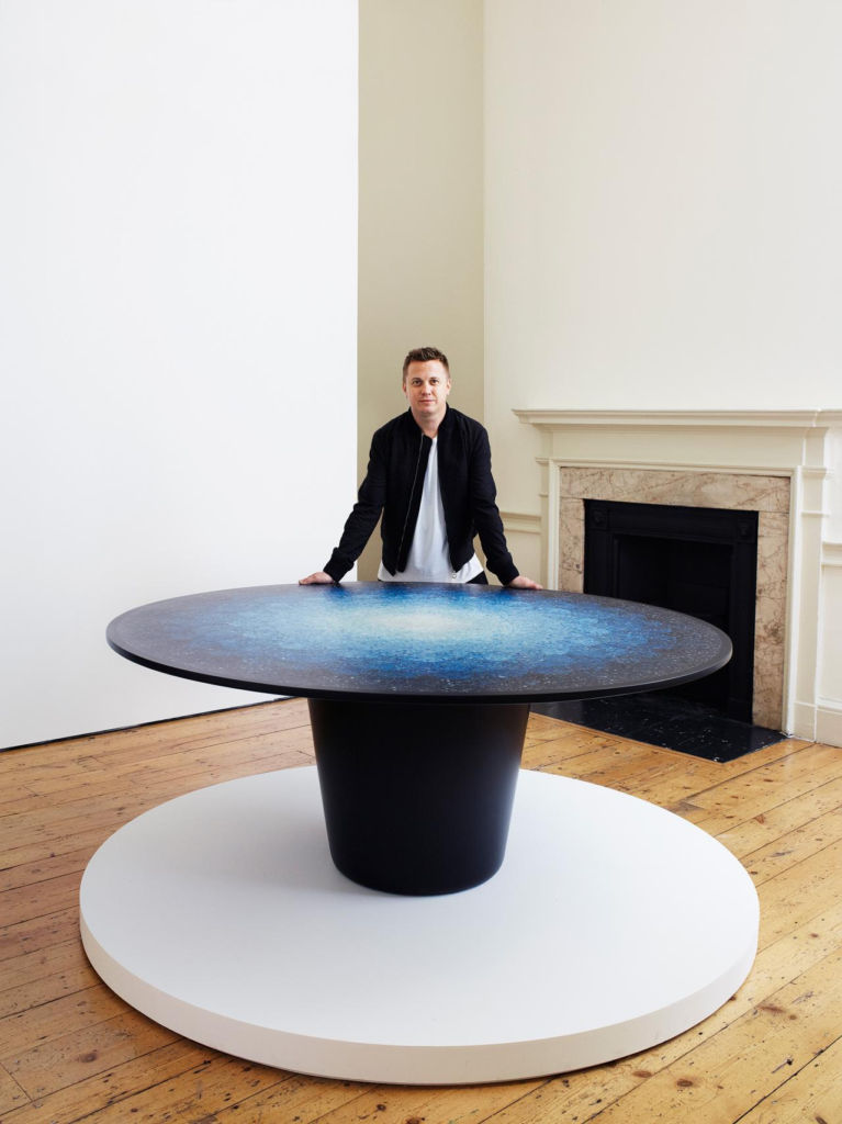 Brodie Neill with his Gyro table - a circular tabletop with a cylindrical base. The tabletop is an ombre terrazzo-like pattern made of micro-plastics from the ocean