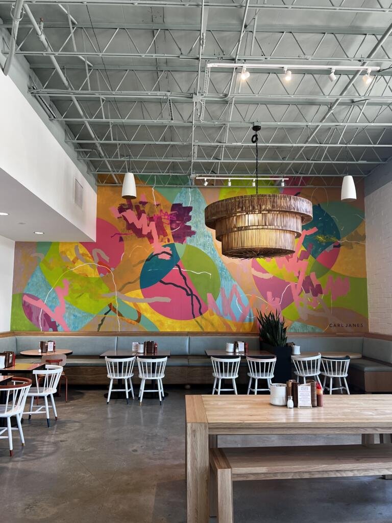 Instagrammable view ofThe Daily West Midtown with a large, colorful mural, modern boho oversized light fixture, concrete floors, wood dining table and bench, and light sage blue/green cushions lining the seats