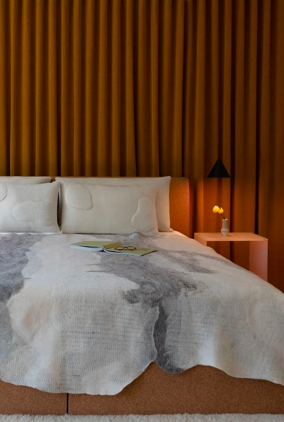 bedroom with a compressed golden velvet curtain hanging from wall to wall. Soft warm lighting, natural bedspread and a minimalist salmon colored nightstand